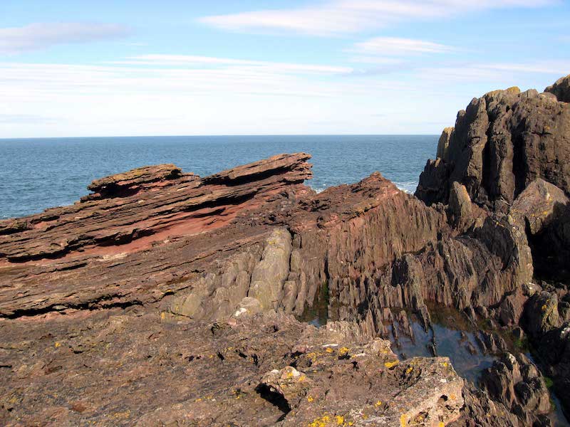 Figure 5: Photograph of Siccar Point showing vertically oriented gray rocks and tilted red sandstone layers.